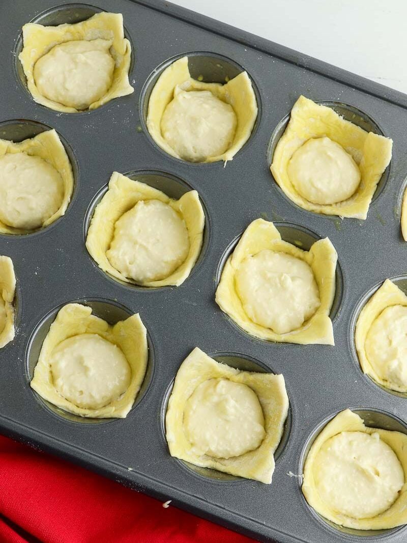 Unbaked mini pies in a muffin tin, each lined with pastry dough and filled with a creamy filling, set on a white surface with a red cloth beside it.
