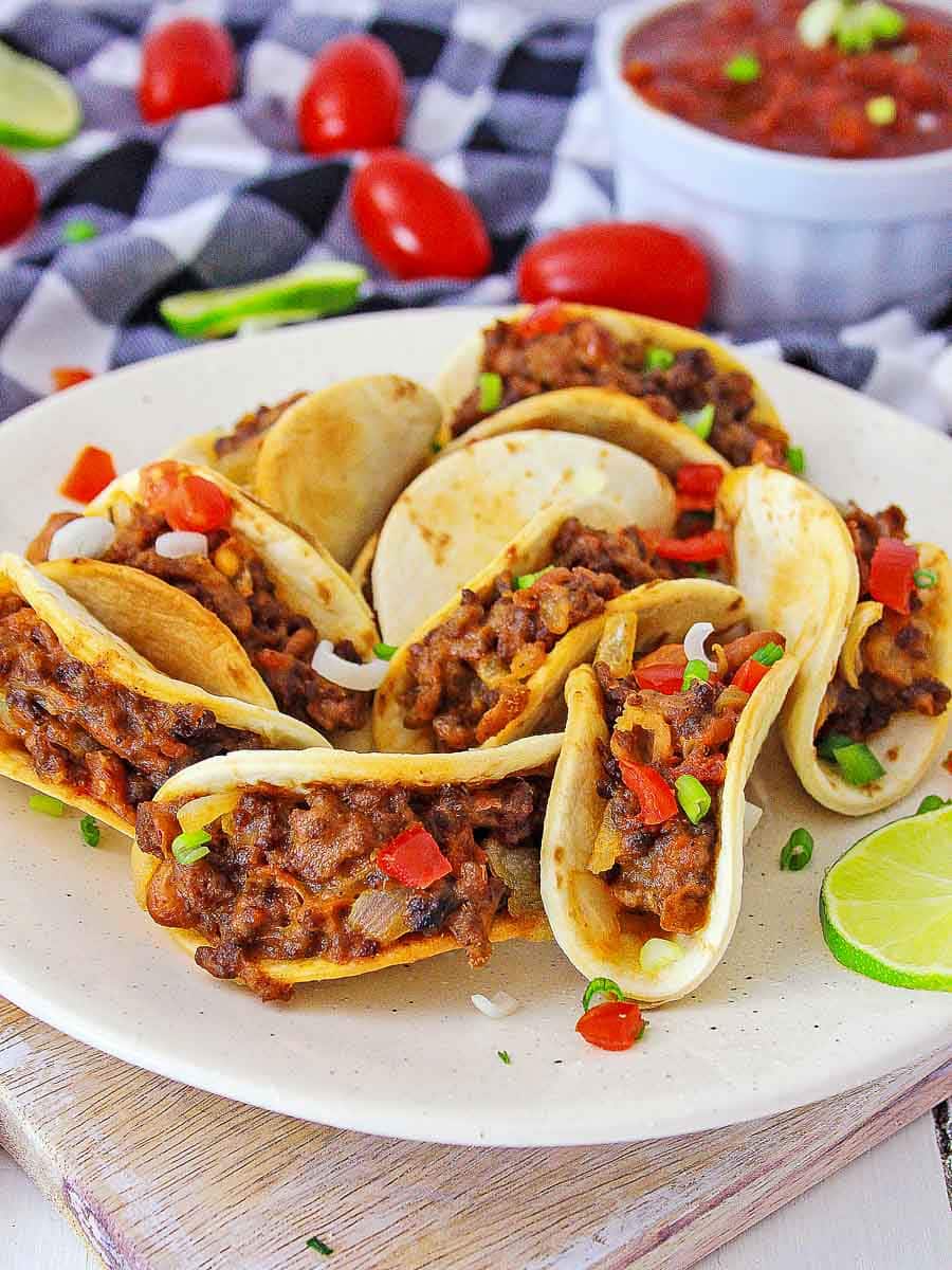 Four tacos with ground meat and diced vegetables in corn tortillas on a white plate, garnished with lime and served with salsa on a vibrant background.