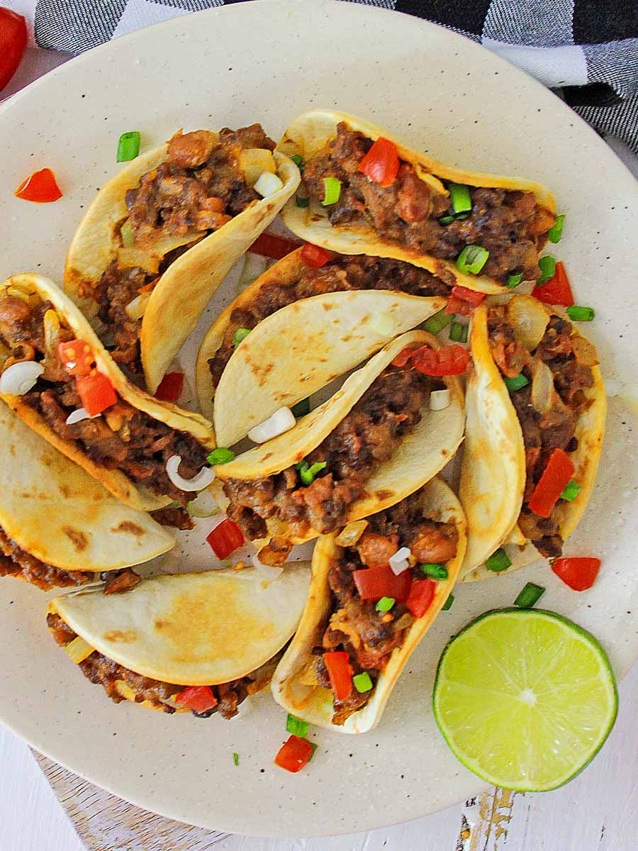 A plate of tacos with ground meat filling, garnished with diced red and green peppers, served with a lime wedge on the side.