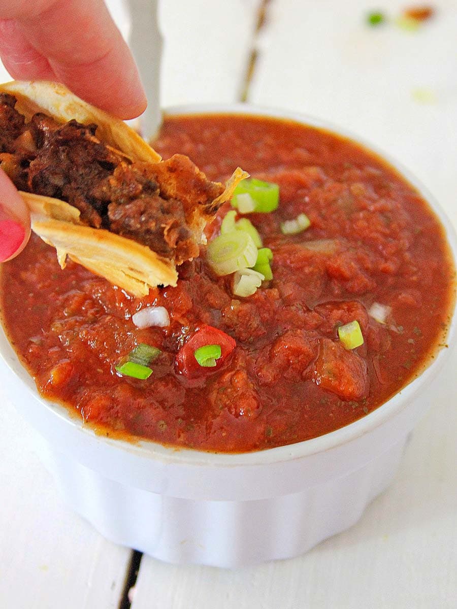 A hand dipping a mini tacos into a bowl of chunky chili garnished with diced scallions.
