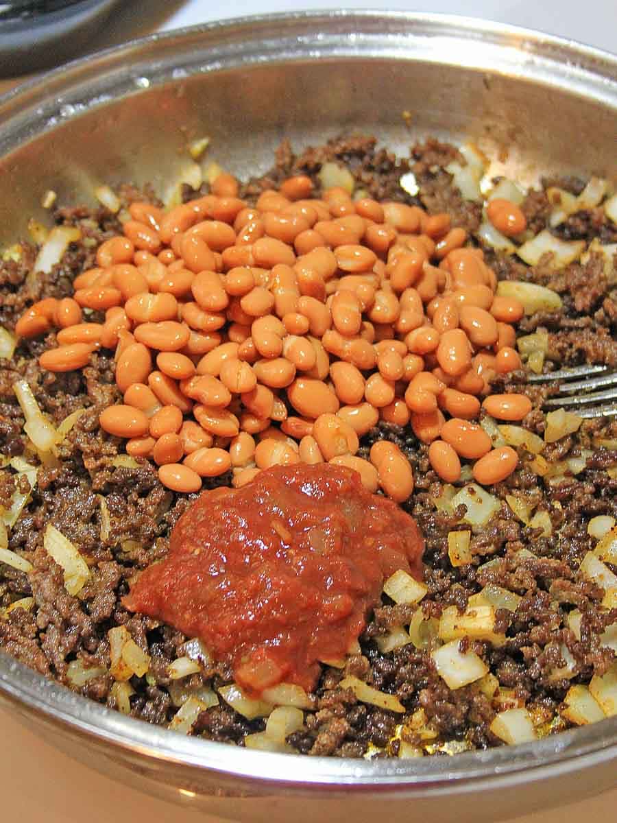 Cooked ground beef with diced onions in a metal skillet, topped with baked beans and a dollop of tomato sauce.