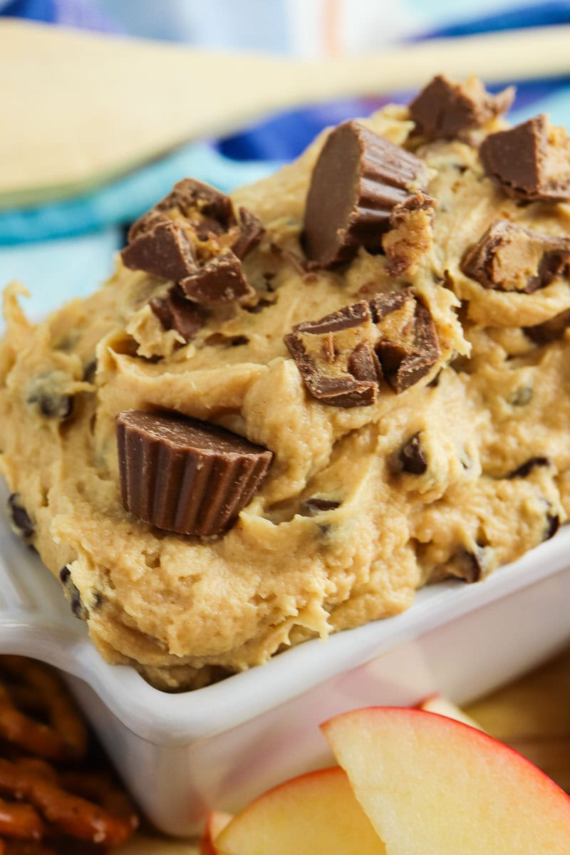 A bowl of peanut butter dip topped with chopped peanut butter cups, surrounded by sliced apples and pretzels on a wooden board.