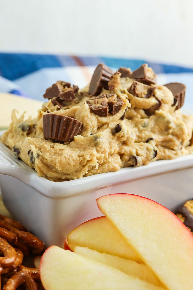 A dish of peanut butter dip topped with chocolate pieces served with apple slices and pretzels on the side.
