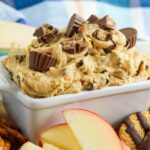 A dish of peanut butter dip topped with chocolate pieces served with apple slices and pretzels on the side.