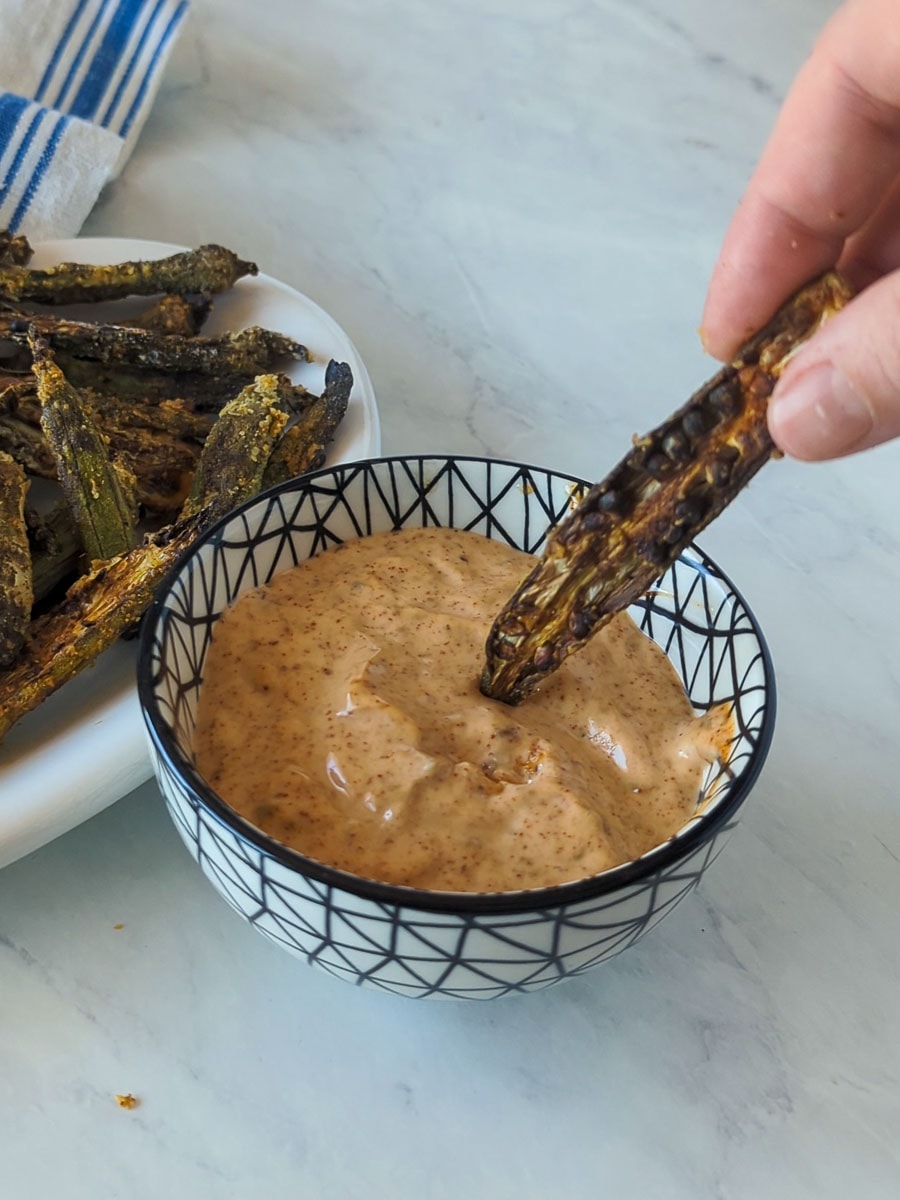 A hand dipping a piece of roasted okra into a bowl of creamy dipping sauce next to a plate of more roasted okra on a marble surface.