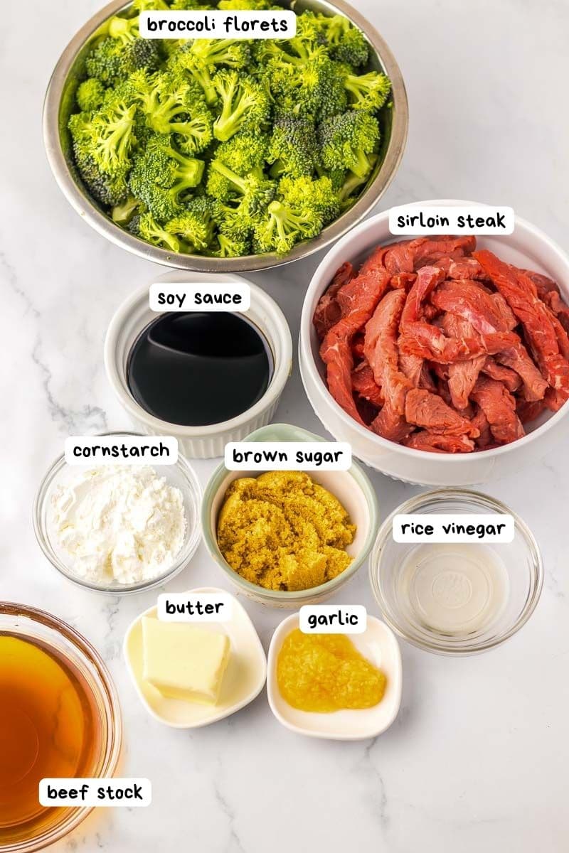 A top-down view of ingredients for a beef and broccoli dish, including bowls of broccoli florets, sliced sirloin steak, soy sauce, cornstarch, brown sugar, rice vinegar, butter, garlic, and beef stock.