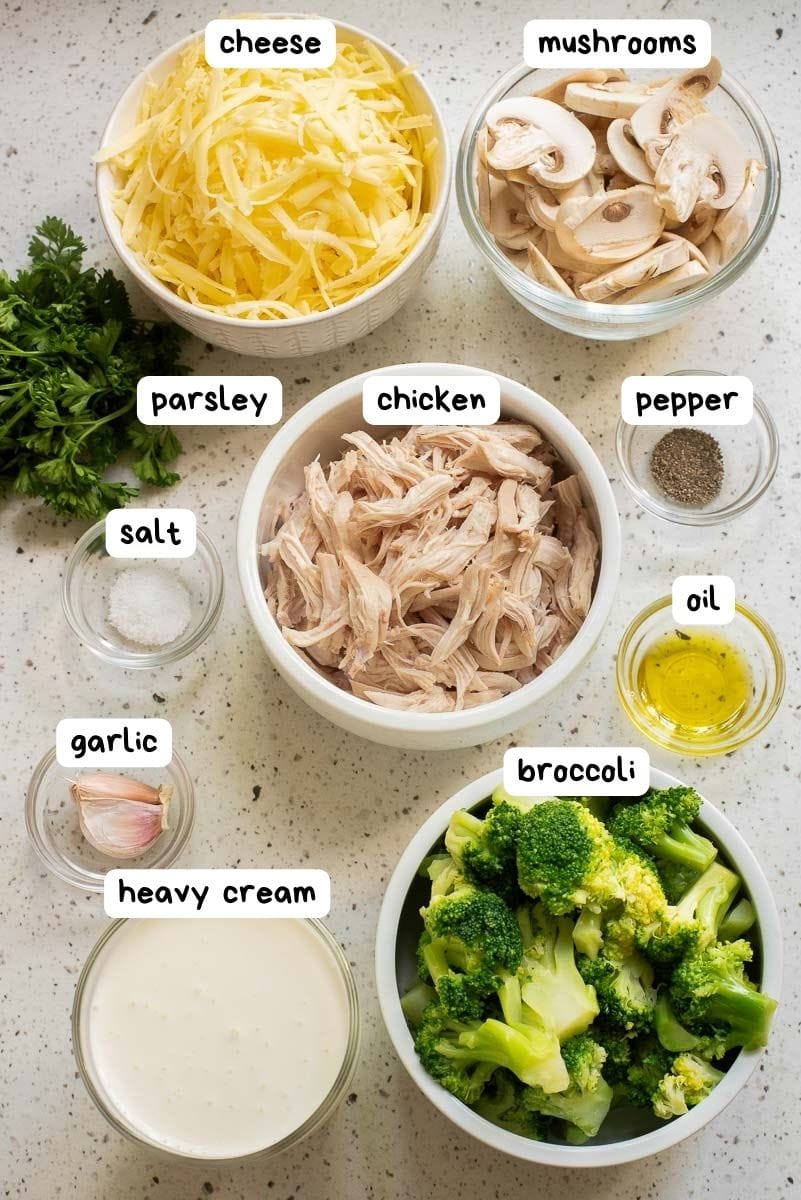 Ingredients arranged on a countertop: shredded cheese, sliced mushrooms, parsley, shredded chicken, black pepper, salt, oil, minced garlic, heavy cream, and broccoli florets.