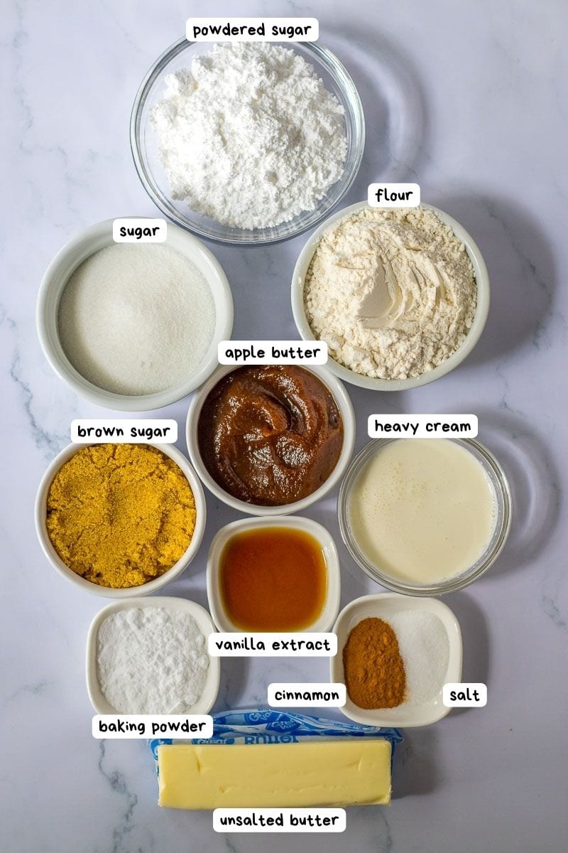 Ingredients on a marble countertop: powdered sugar, flour, sugar, brown sugar, apple butter, heavy cream, baking powder, vanilla extract, cinnamon, salt, and a stick of unsalted butter.