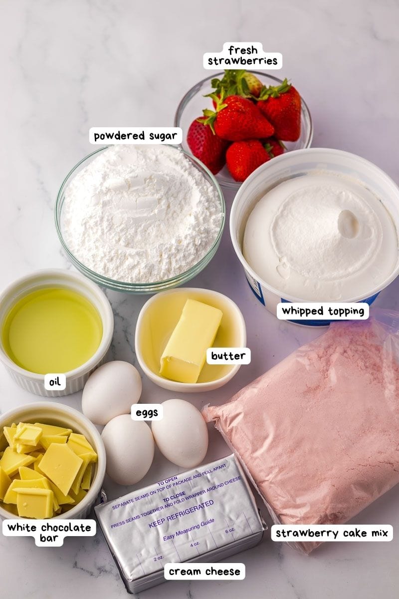 Various baking ingredients are spread on a white surface, including fresh strawberries, powdered sugar, whipped topping, oil, butter, eggs, white chocolate bar, cream cheese, and strawberry cake mix.