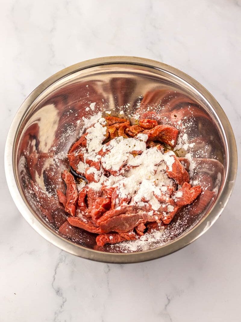 A stainless steel bowl filled with slices of raw beef, lightly coated with cornstarch, on a white marble surface.
