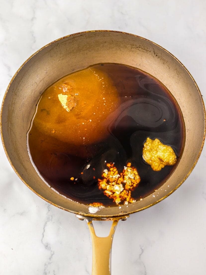 A frying pan contains oil, soy sauce, minced garlic, and possibly ginger on a marble surface, appearing to be the start of a cooking process.