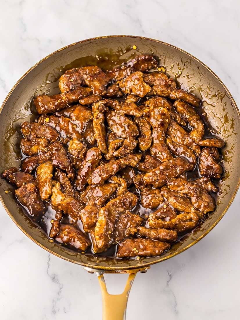 A skillet filled with cooked beef strips coated in a brown sauce, placed on a marble countertop.