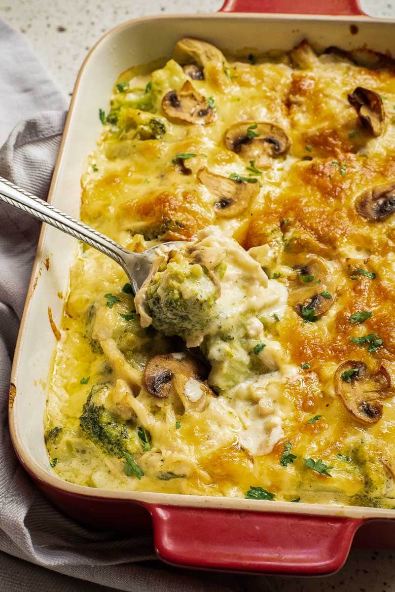 A spoon lifts a portion of a cheesy casserole with broccoli, mushrooms, and a creamy sauce from a red baking dish.