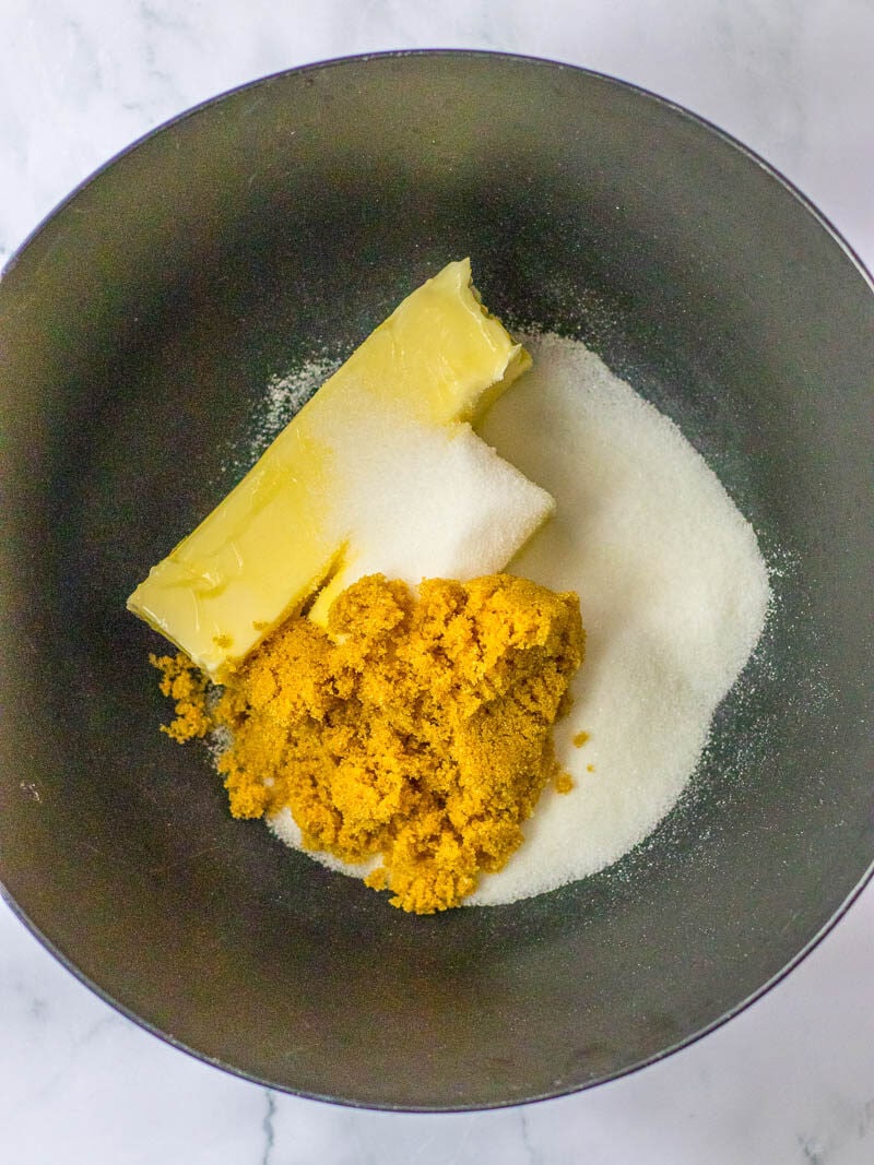 A black bowl contains a stick of butter, a pile of brown sugar, and a pile of granulated sugar on a white marble background.