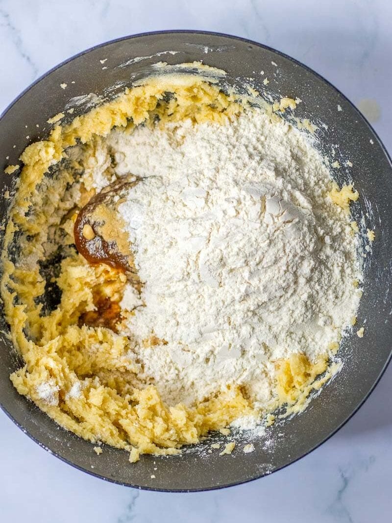A mixing bowl with a creamy yellow batter and a heap of flour on top, ready to be mixed. The bowl sits on a white marble surface.
