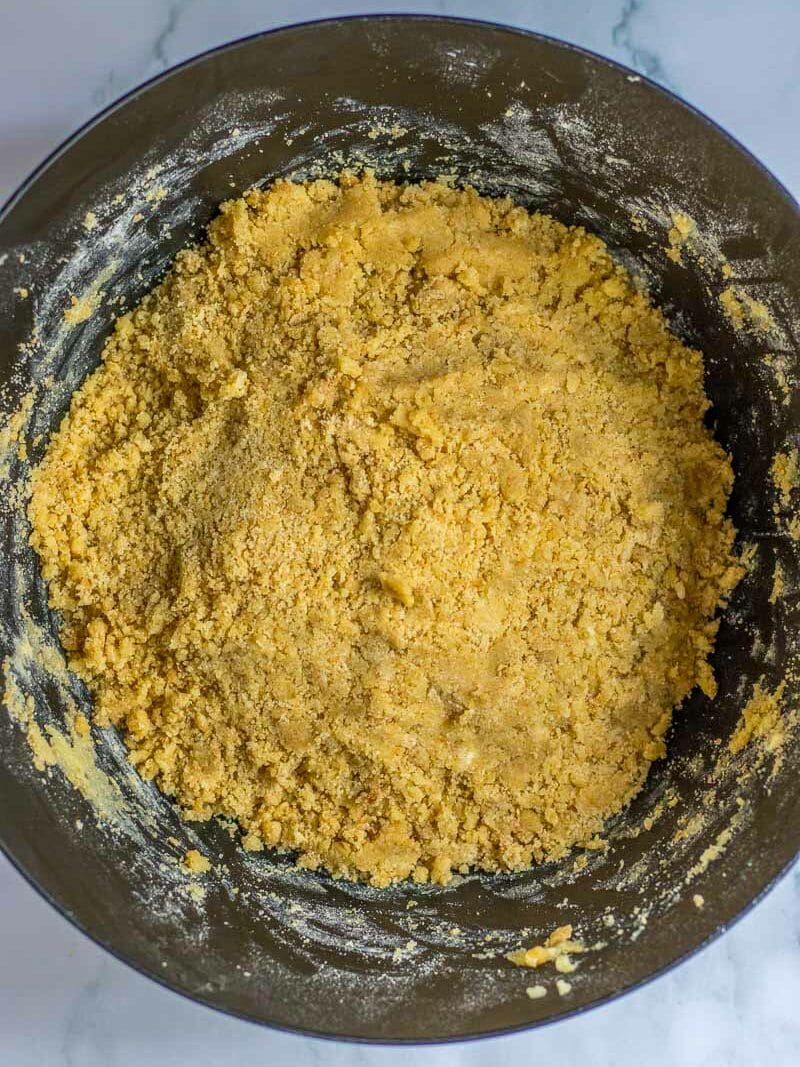 Close-up view of a mixing bowl filled with crumbly, yellow dough being prepared on a marble surface.
