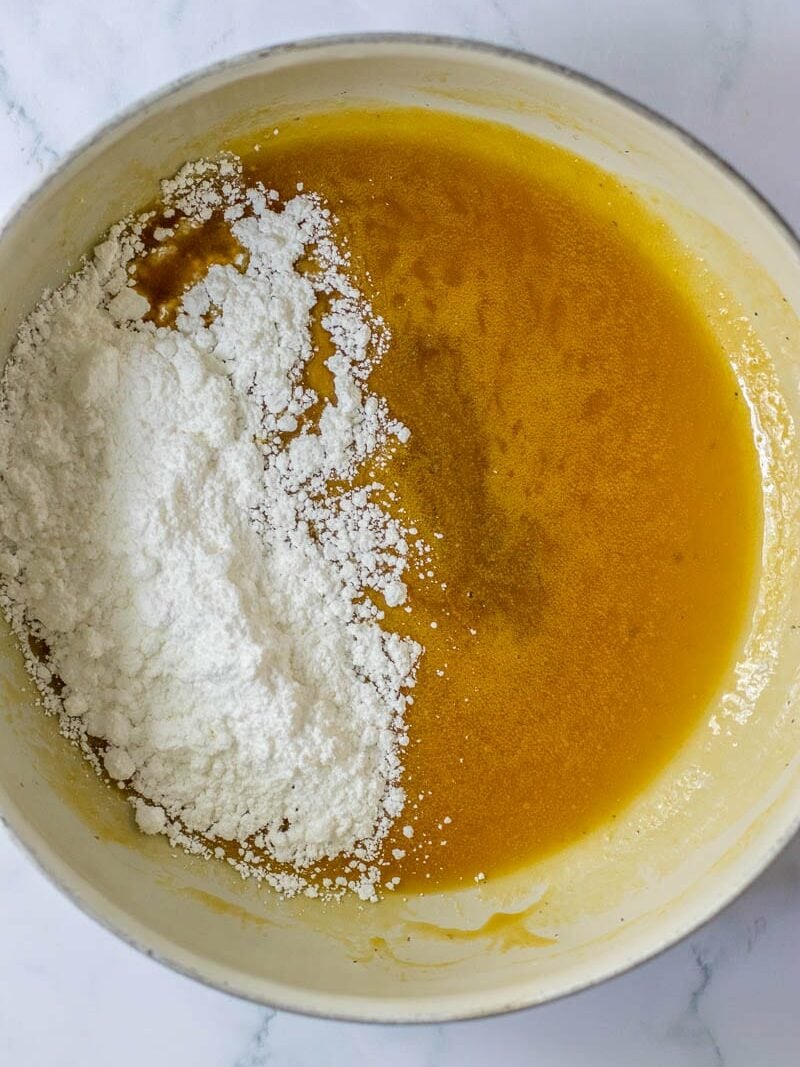 A mixing bowl containing a yellow liquid mixture and a portion of white flour added on the left side.