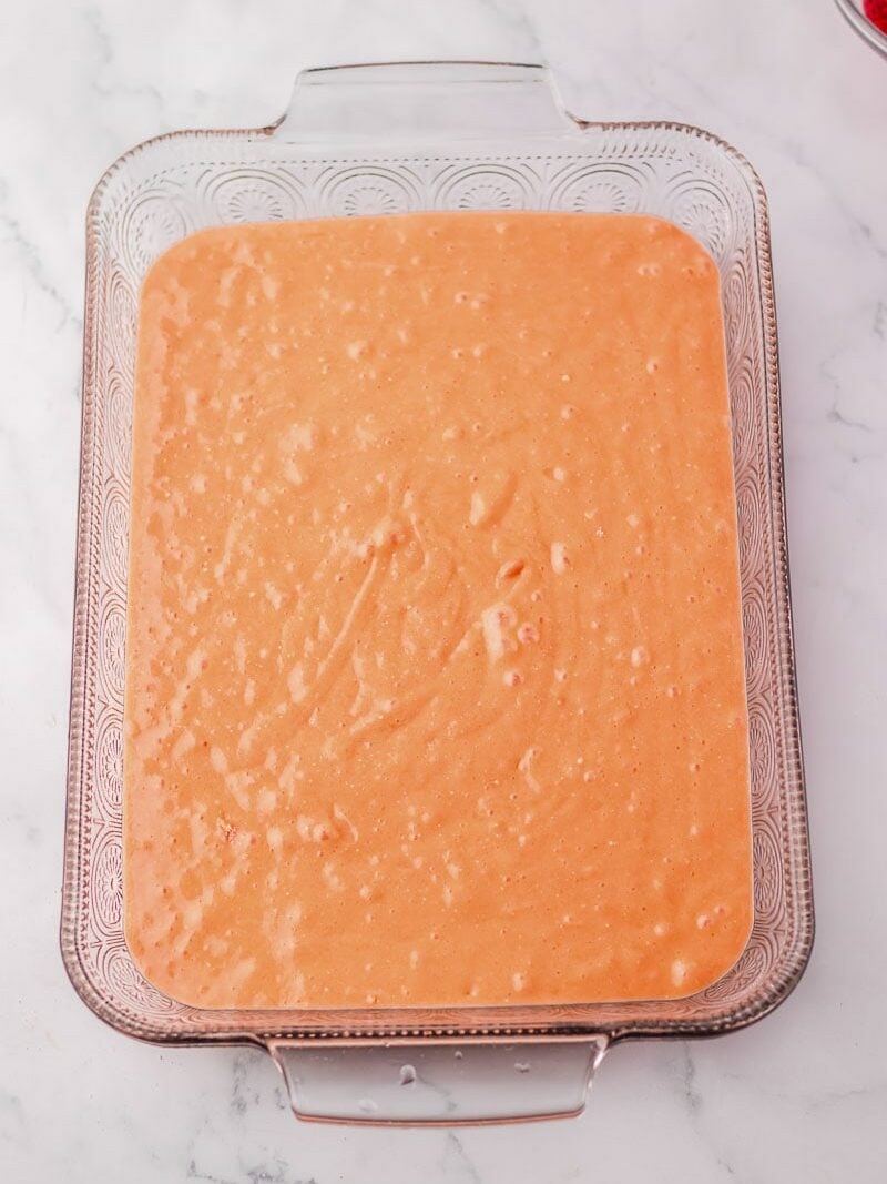 A rectangular glass baking dish filled with an unbaked strawberry cake sits on a white marble surface. A small bowl with strawberries is partially visible in the corner.