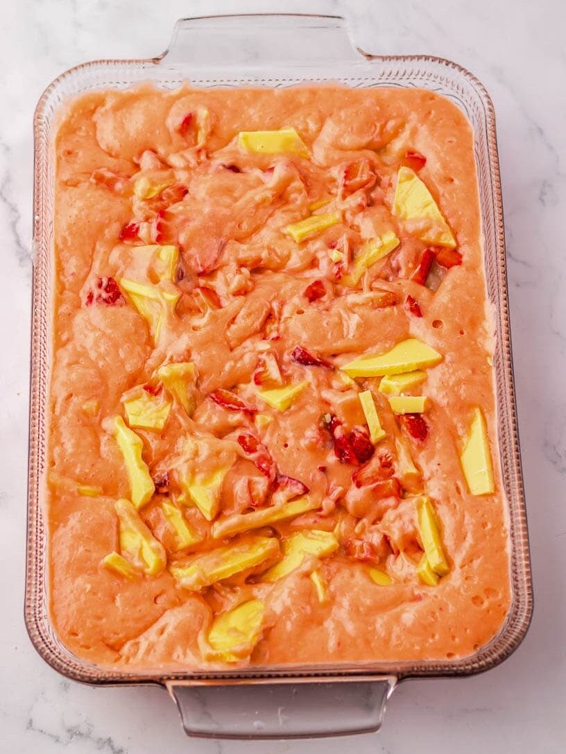 A rectangular glass baking dish filled with an unbaked, strawberry cake batter, topped with chunks of white chocolate and strawberries.