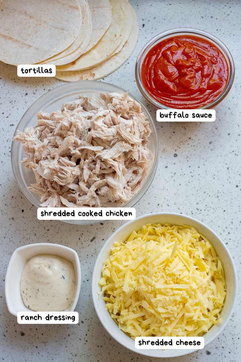 An overhead view of labeled ingredients: tortillas, buffalo sauce, shredded cooked chicken, ranch dressing, and shredded cheese, arranged in bowls on a speckled countertop.
