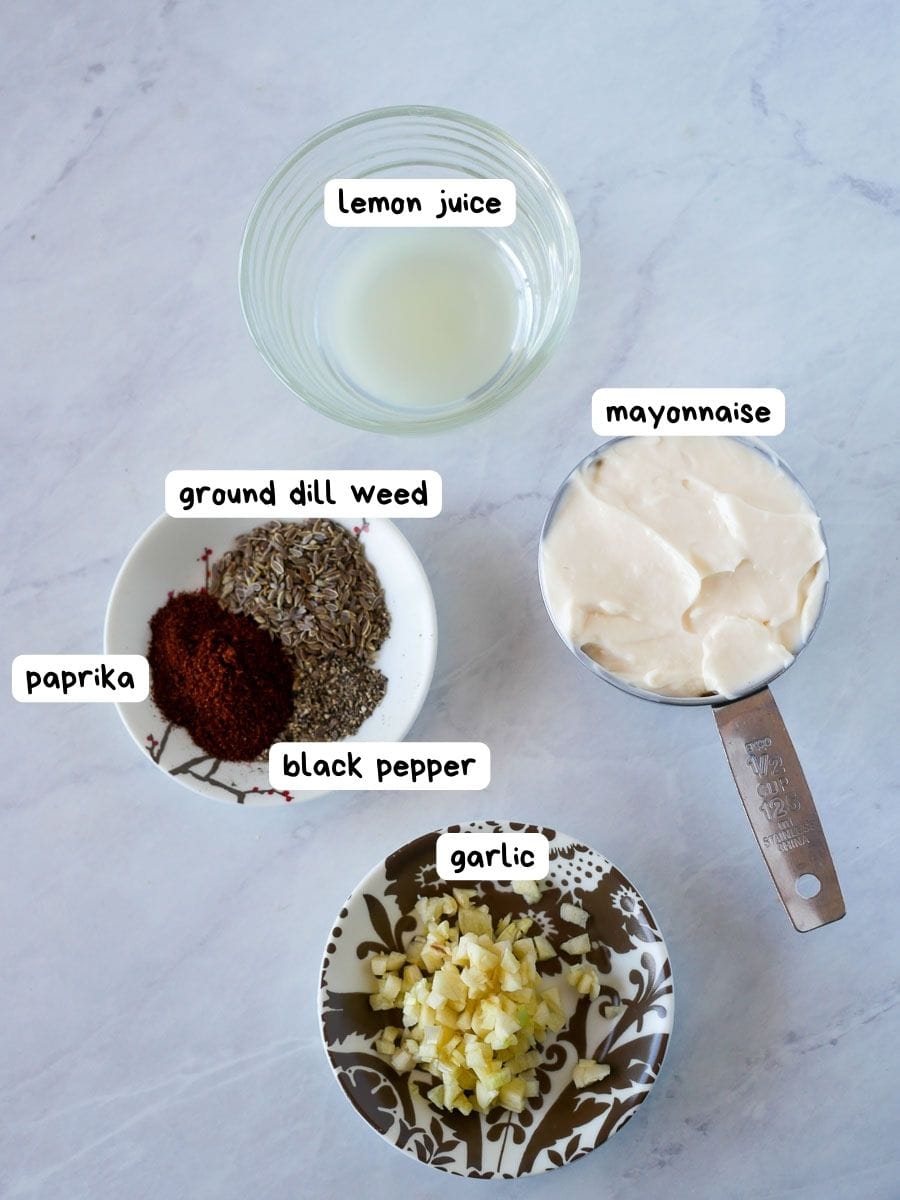 Ingredients laid out on a marble surface: a glass bowl of lemon juice, a measuring cup of mayonnaise, and small bowls containing paprika, ground dill weed, black pepper, and chopped garlic.