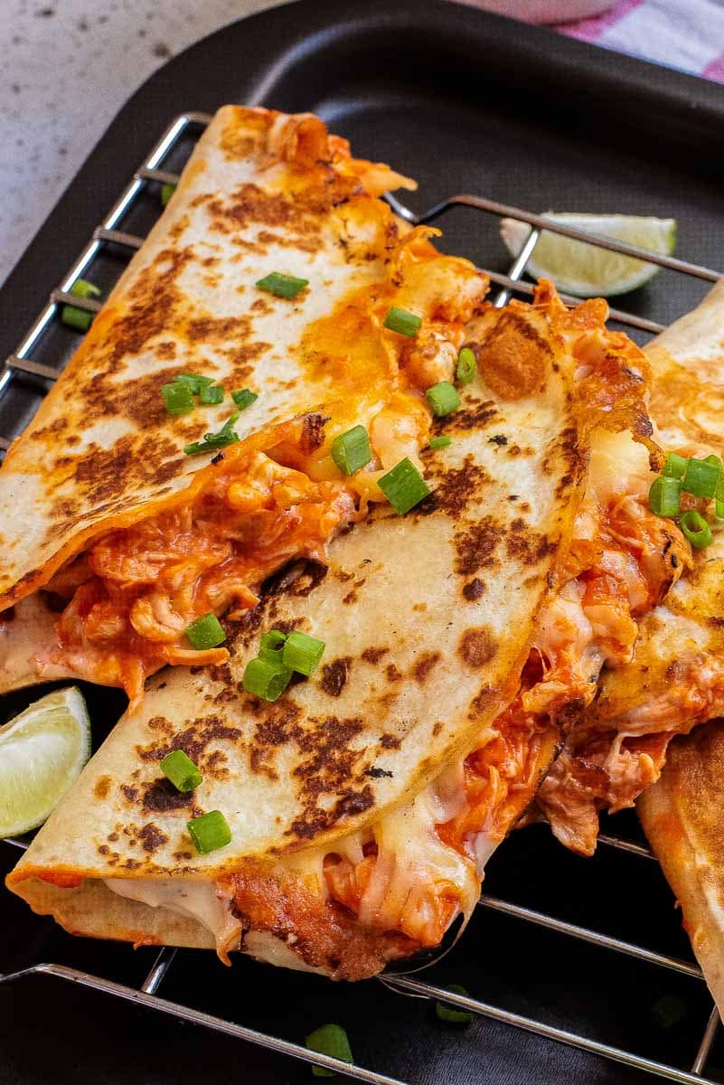 Close-up of two grilled quesadillas filled with melted cheese and chicken, garnished with chopped green onions, served on a metal rack with lime wedges in the background.