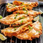Four chicken quesadillas garnished with chopped green onions and served with lime wedges on a wire rack.