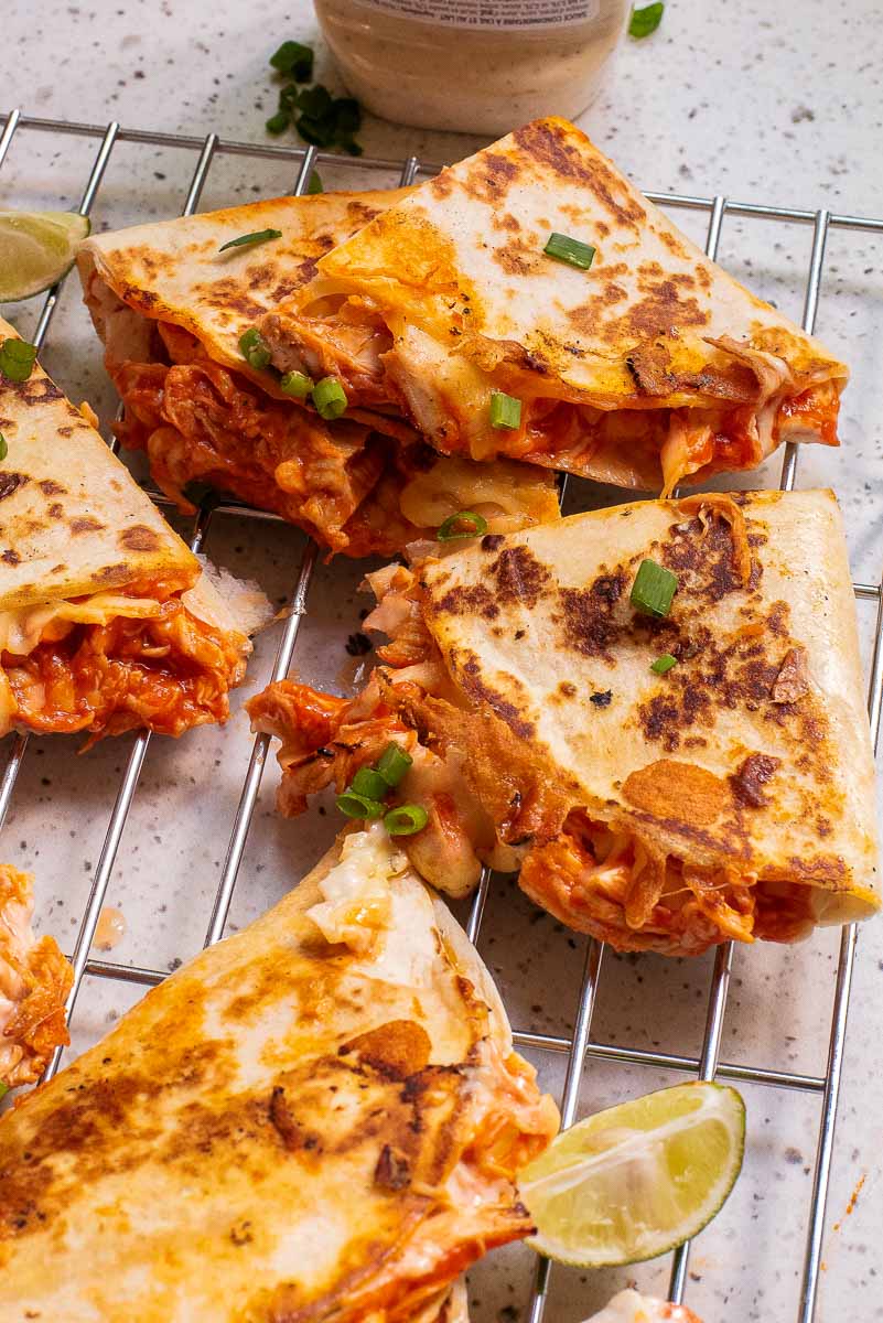 Five grilled quesadilla slices filled with cheese and shredded chicken, garnished with green onions, are placed on a wire rack. Lime wedges are visible beside the quesadillas.