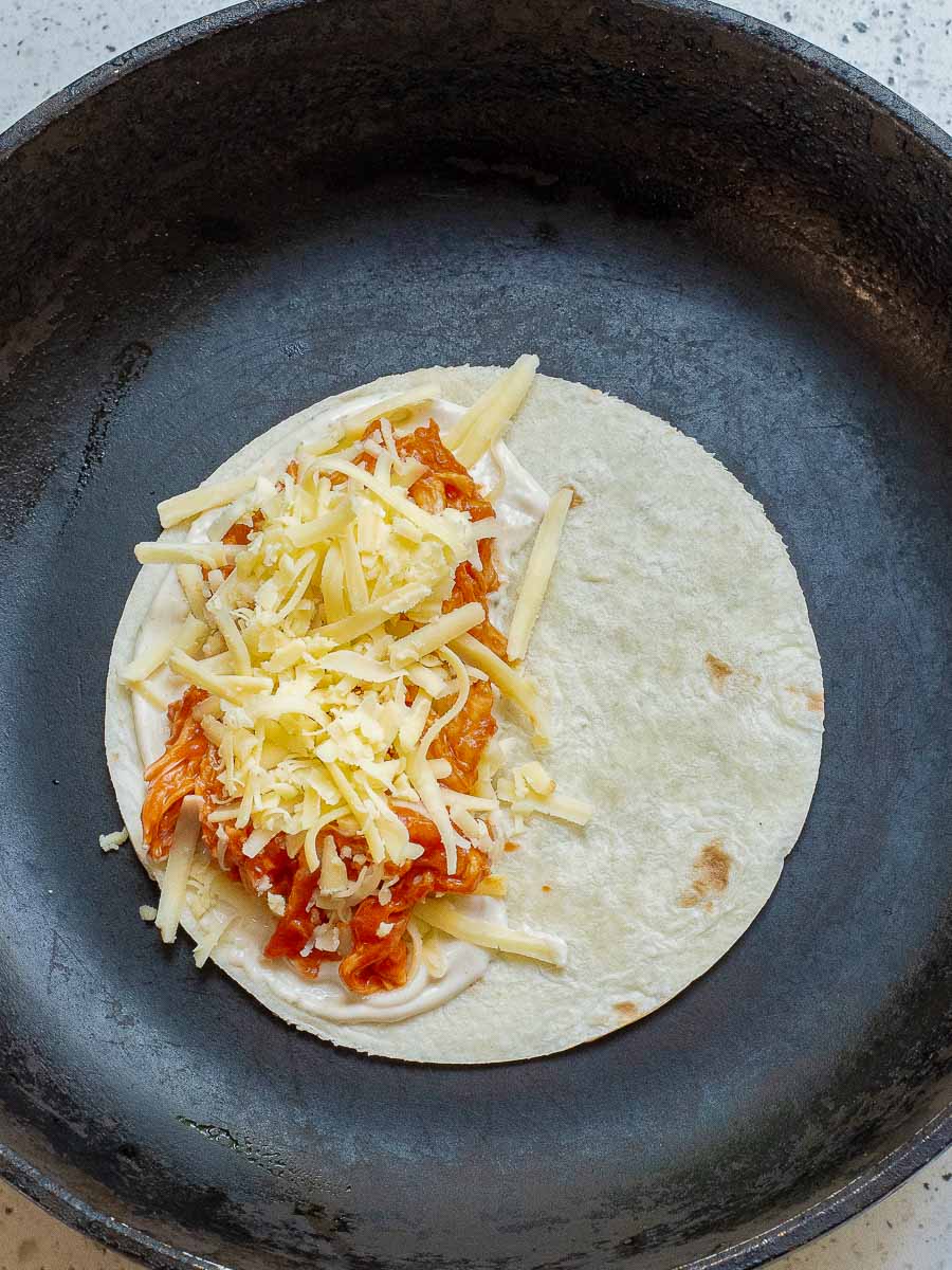 A tortilla with shredded chicken, cheese, and sauce on top, placed in a black skillet, ready for cooking.