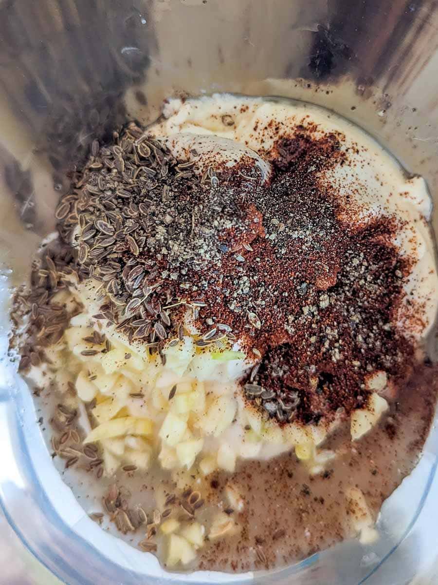 A blender jar containing yogurt, chopped garlic, cumin seeds, ground black pepper, and red chili powder.