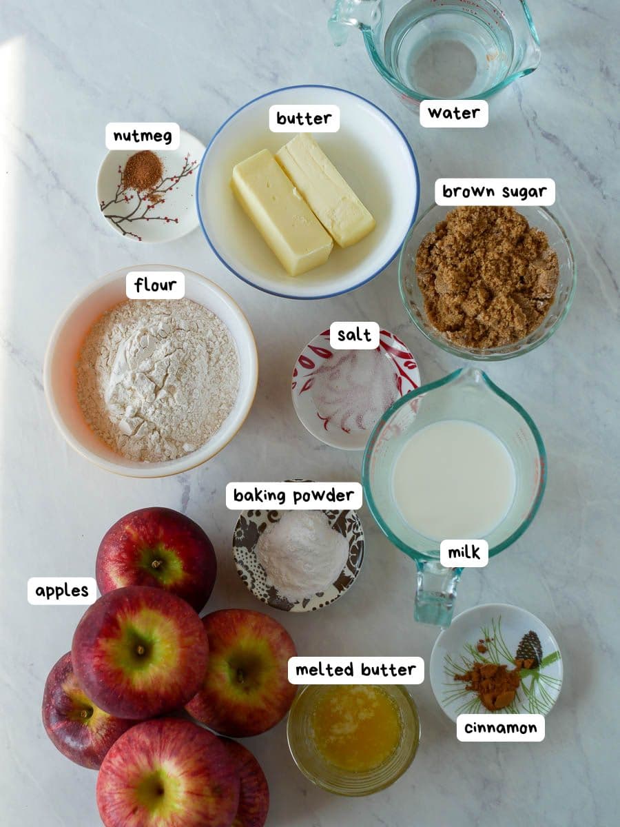 Top-down view of neatly arranged apple cinnamon bake ingredients: apples, flour, butter, water, brown sugar, salt, baking powder, milk, melted butter, nutmeg, and cinnamon on a marble surface.
