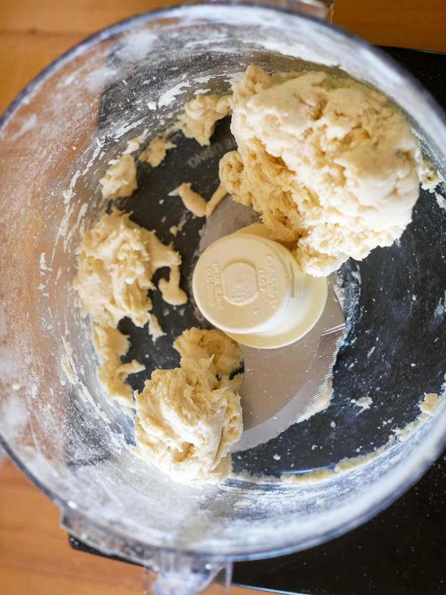 A food processor bowl with partially mixed dough forming several clumps on the bottom.