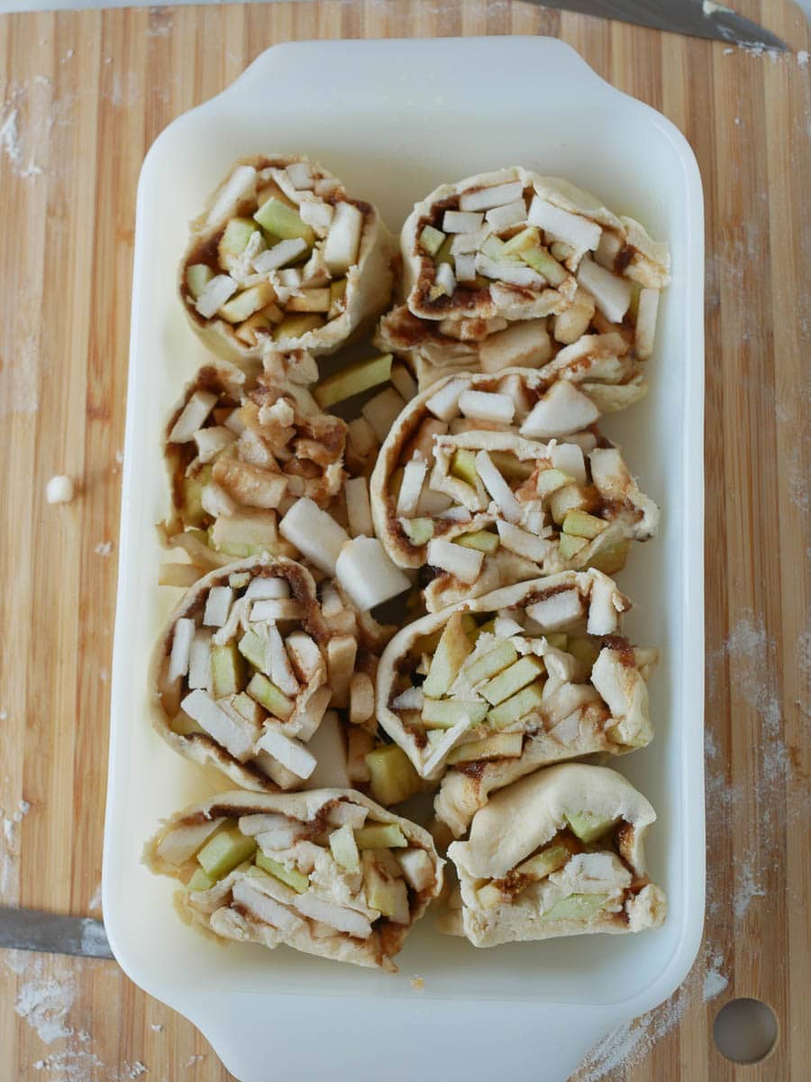 Unbaked apple and pear pastry rolls in a white baking dish on a wooden surface.