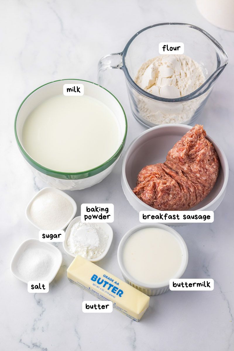 Ingredients for a recipe are arranged on a counter: flour, breakfast sausage, milk, sugar, baking powder, salt, butter, and buttermilk, each in separate bowls or containers.