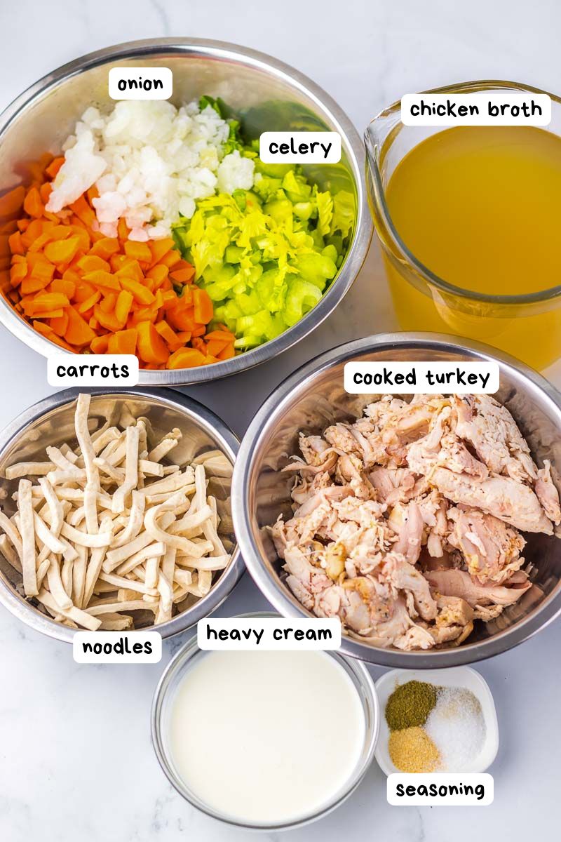 Ingredients for turkey soup: chopped onion, celery, carrots, cooked turkey, chicken broth, noodles, heavy cream, and seasoning, all labeled in separate bowls on a counter.
