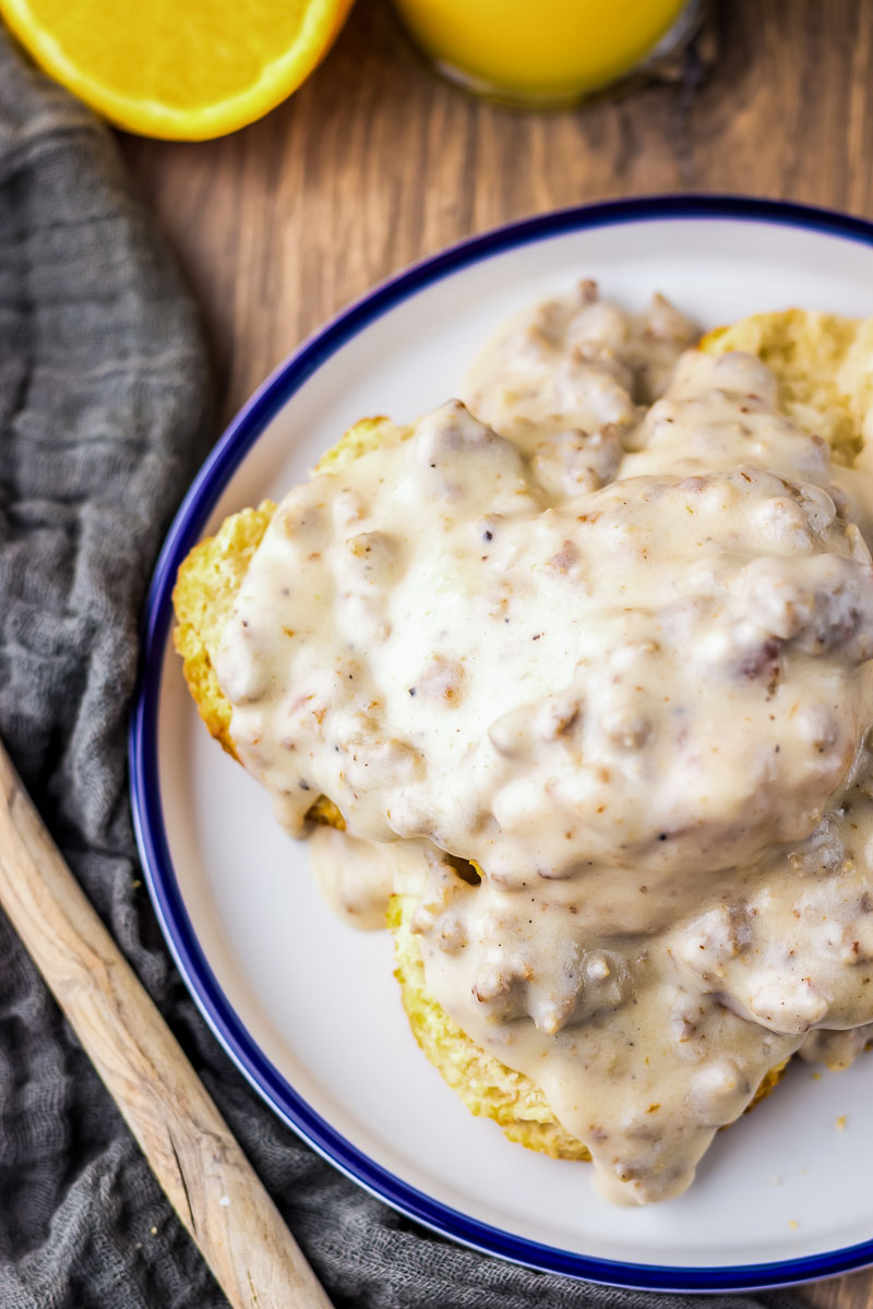 Biscuits topped with thick sausage gravy on a white plate, with a slice of orange and a gray cloth nearby.