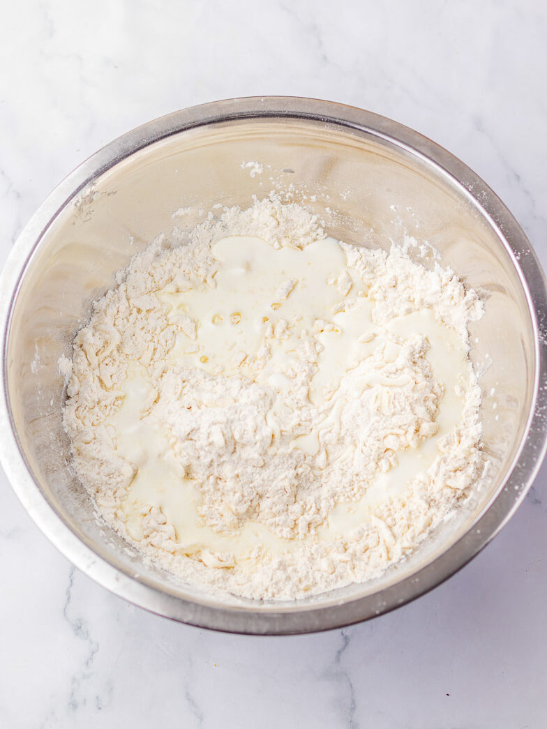 A stainless steel bowl contains flour and milk being mixed on a marble countertop.