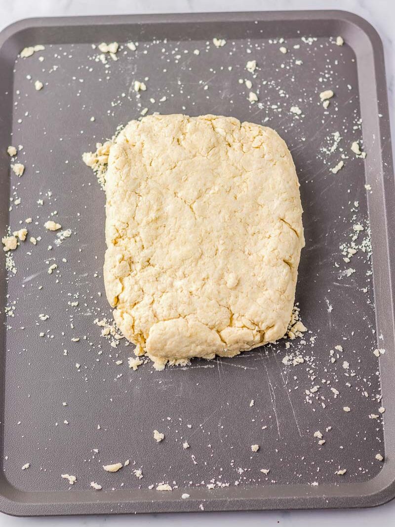 A rectangular, rough block of dough rests on a baking sheet, surrounded by scattered crumbs.