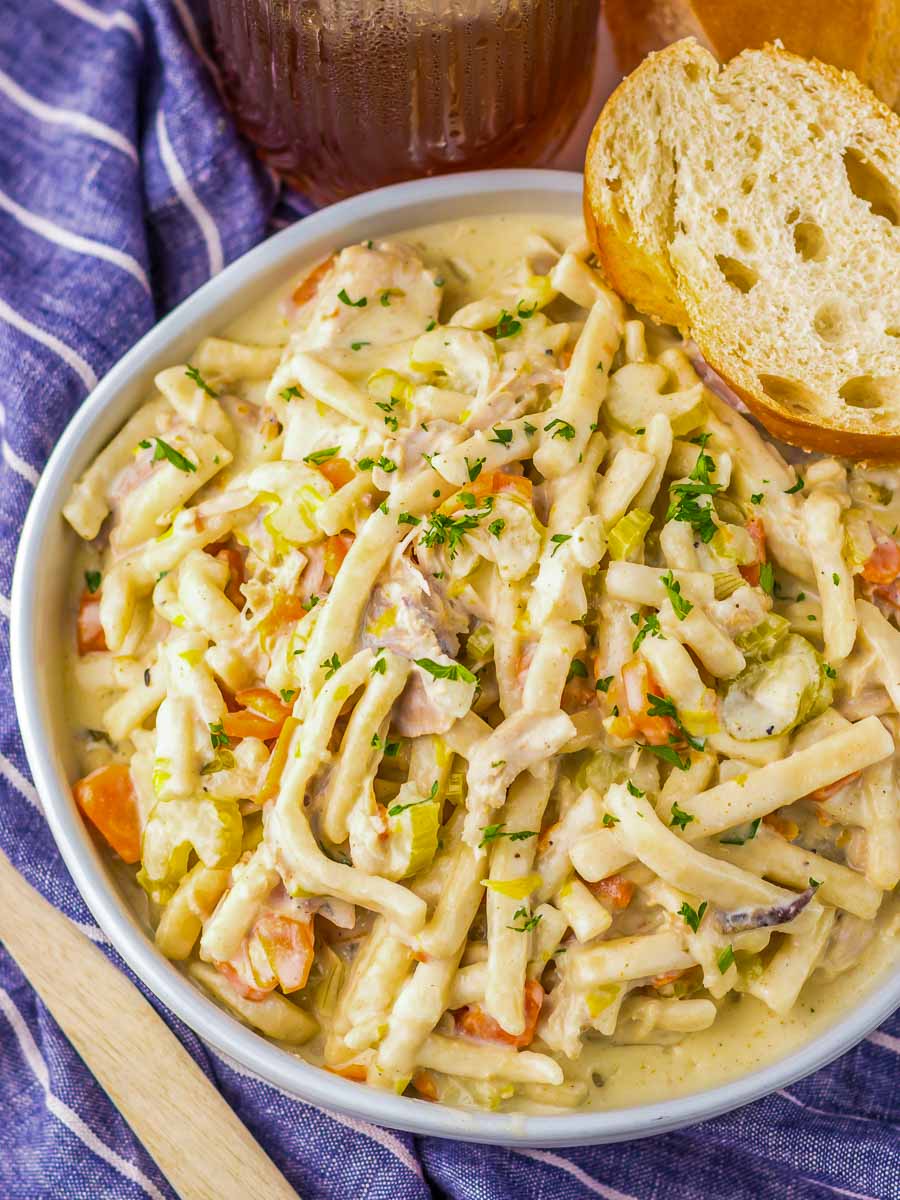 A bowl of creamy chicken noodle casserole topped with chopped parsley sits on a table with a piece of bread.