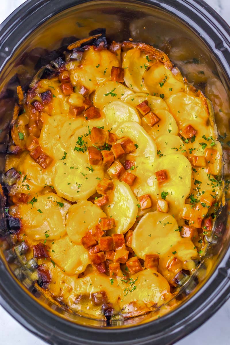 A crockpot filled with layered sliced potatoes, melted cheese, and diced ham, garnished with parsley.