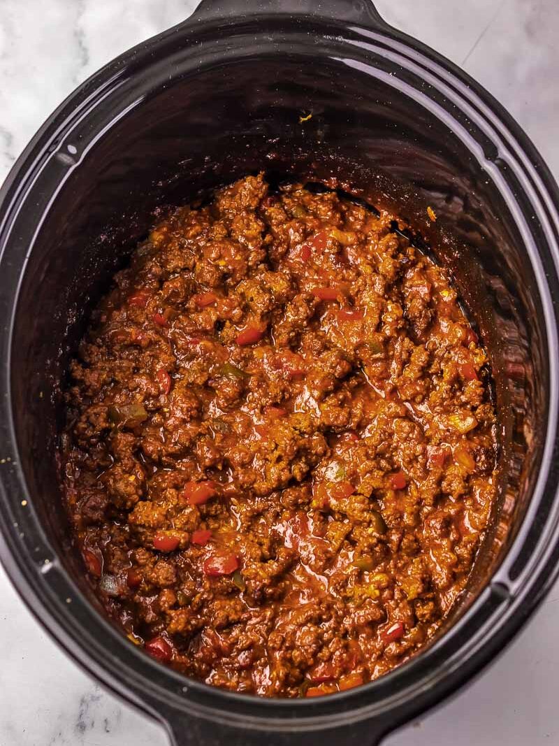 A black slow cooker filled with cooked chili, containing ground meat and diced vegetables in a thick, reddish-brown sauce on a marble countertop.