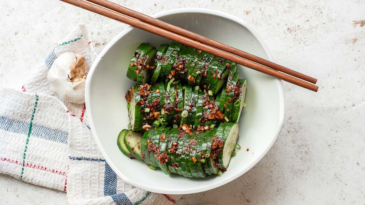 A white bowl of sliced cucumbers topped with chili oil, garlic, and green onions, with a pair of chopsticks resting on the bowl. 