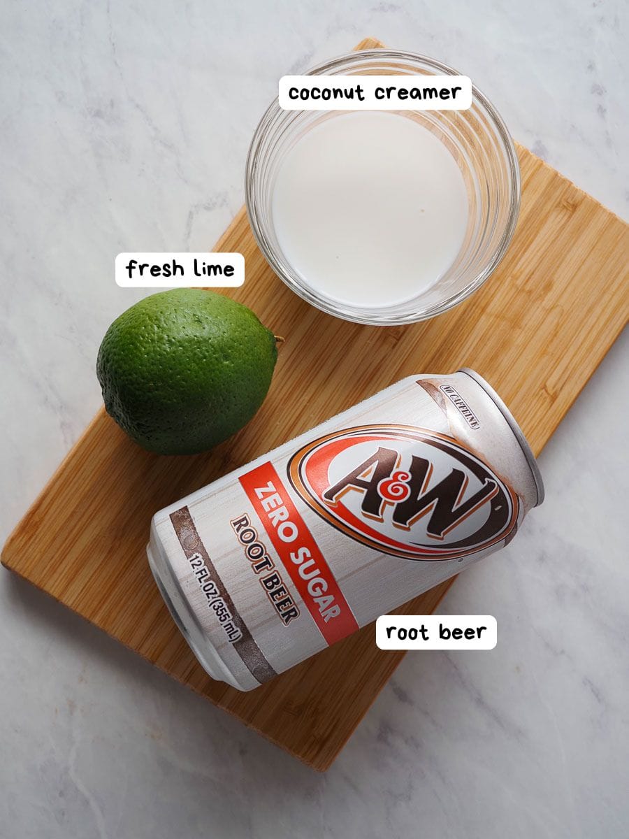 A can of zero sugar root beer, a whole lime, and a glass bowl of coconut creamer on a wooden board, labeled accordingly.