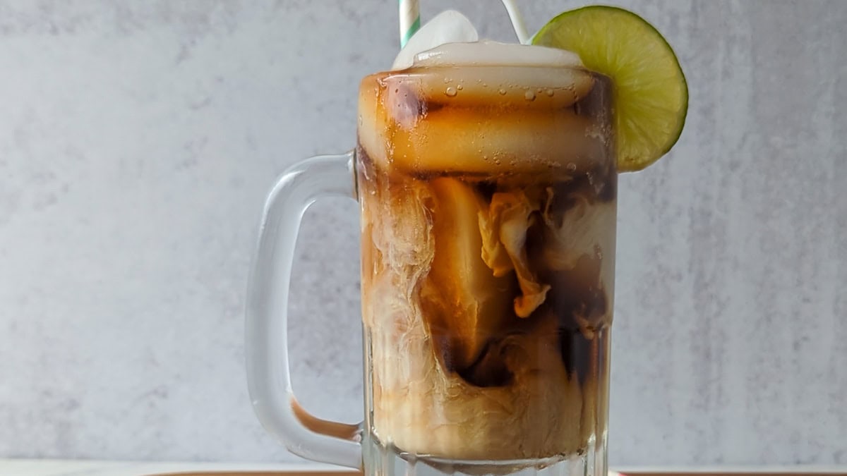 A glass mug filled with iced coffee, topped with foam, ice cubes, a lime slice, and two straws against a light background.