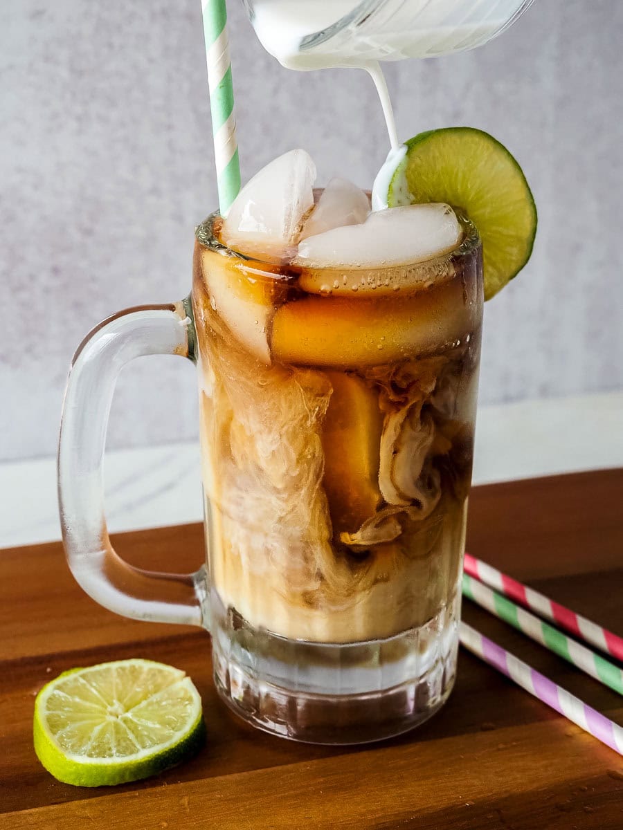 A glass mug filled with iced coffee is topped with milk being poured in, garnished with a lime slice and served with a striped straw; a lime wedge lies nearby.
