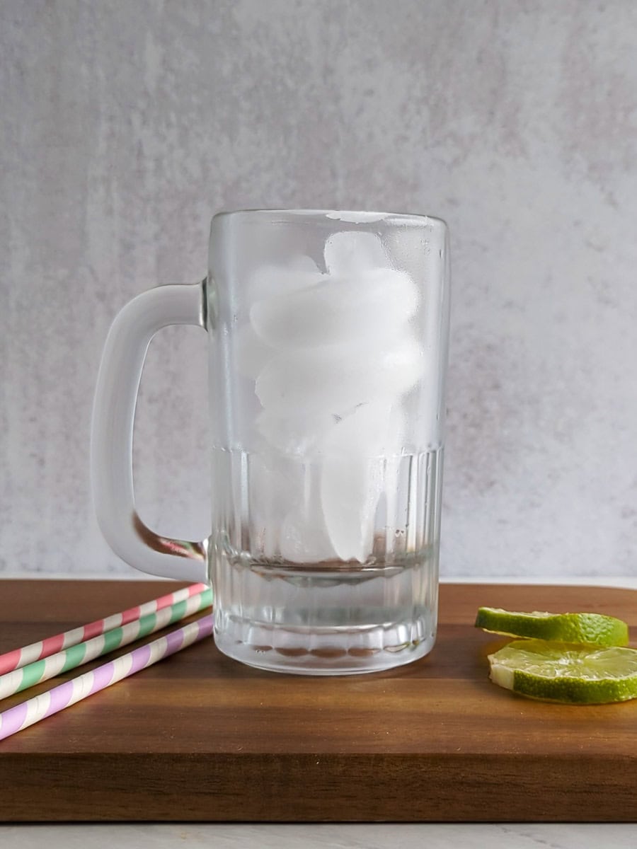 A clear glass mug filled with ice cubes sits on a wooden surface next to two lime wedges and three striped paper straws.