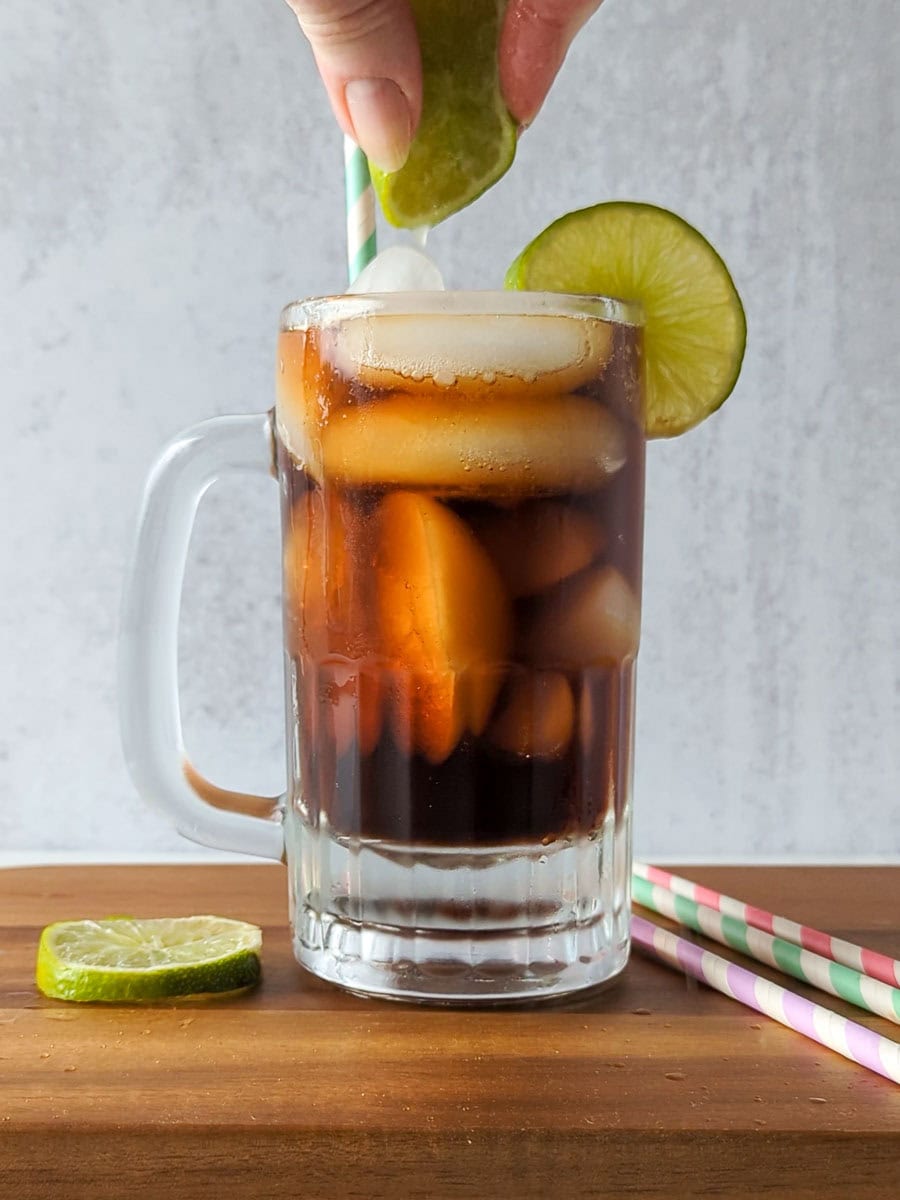 A hand squeezes a lime wedge over a glass mug filled with iced cola, garnished with a lime slice. Striped straws and a lime slice rest on the wooden surface.