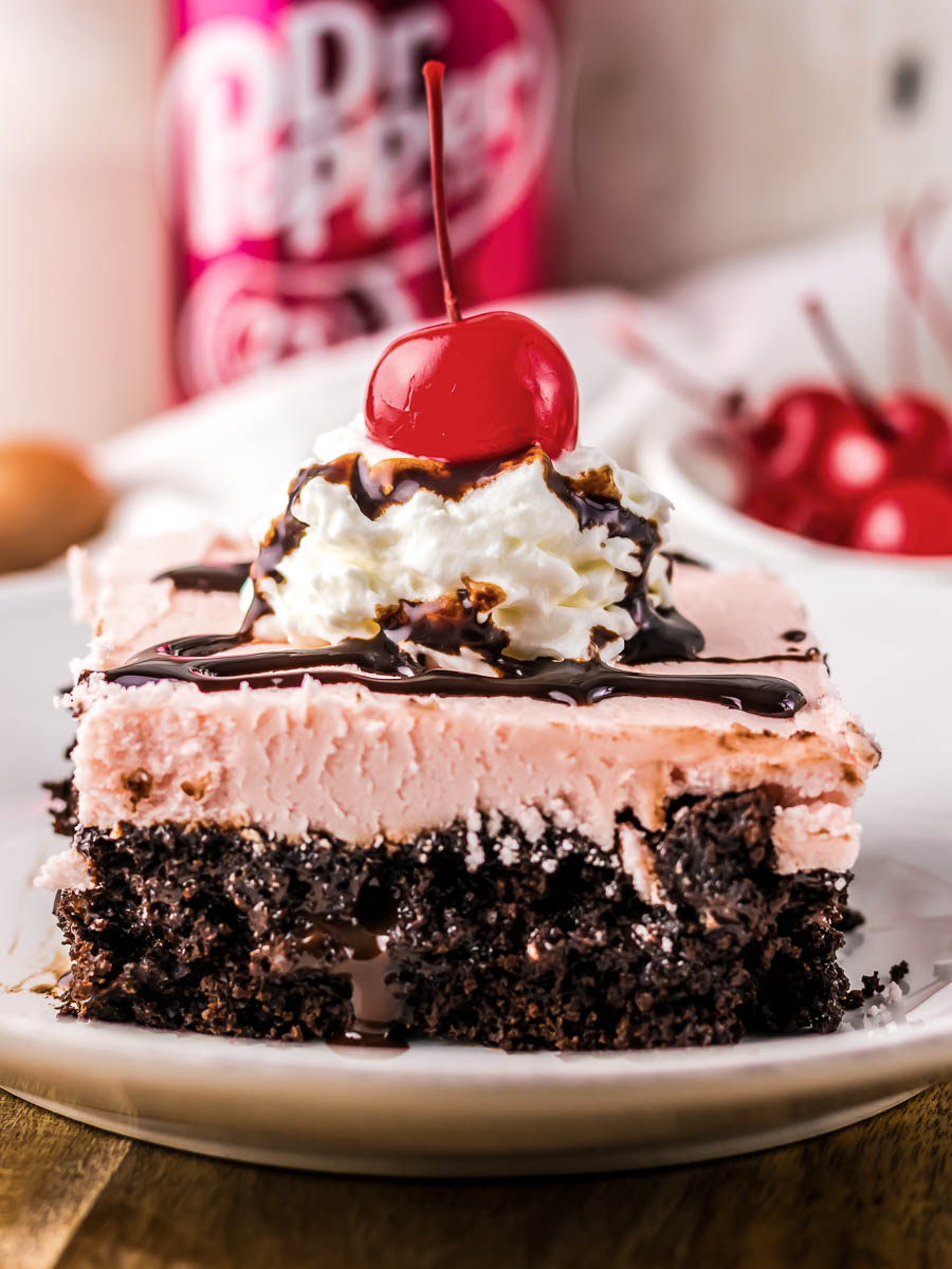 A slice of chocolate and pink layered cake topped with whipped cream, chocolate syrup, and a cherry, on a white plate. A can of soda and more cherries are in the background.