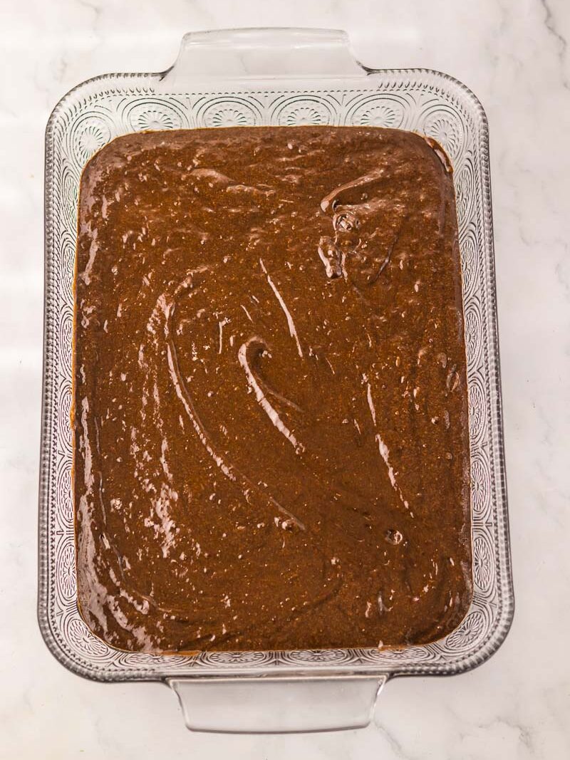 A glass baking dish filled with rich dr pepper cake batter sits on a white marble surface, ready for baking.