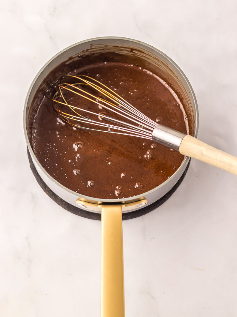 A saucepan filled with rich chocolate mixture for dr pepper cake is being whisked, resting on a marble surface.